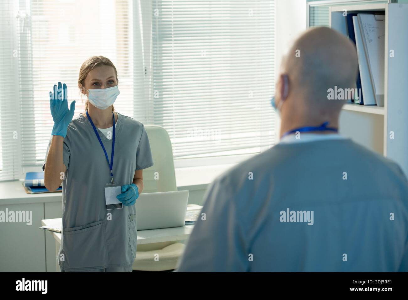 Junge Arbeiterin des medizinischen Labors in schützender Arbeitskleidung winken Hand zu kahlen männlichen Kollegen, während Begrüßung ihn in der Morgen Stockfoto