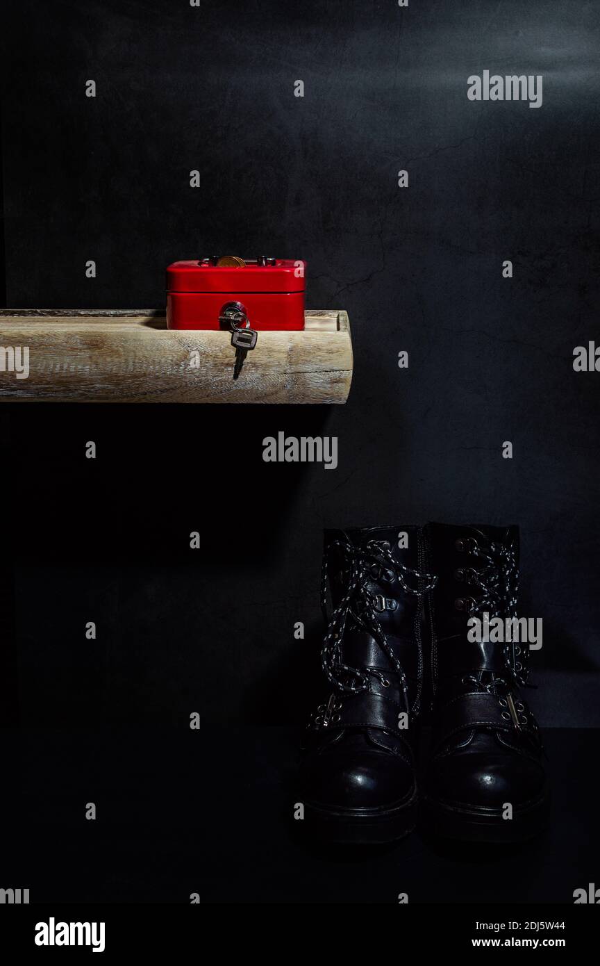 Das Stillleben des roten Geldschatulle auf hölzern Regal vor dunkelgrauem Hintergrund und schwarzen Biker-Stiefeln Auf dem Boden Stockfoto