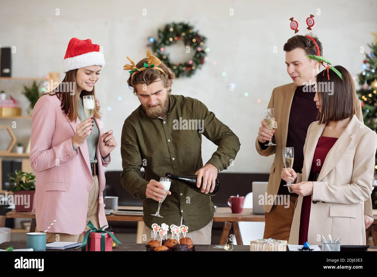 Glücklicher junger bärtiger Geschäftsmann, der Champagner in Flöten von Kollegen gießt Während der Weihnachtsfeier während der Firmenfeier Stockfoto