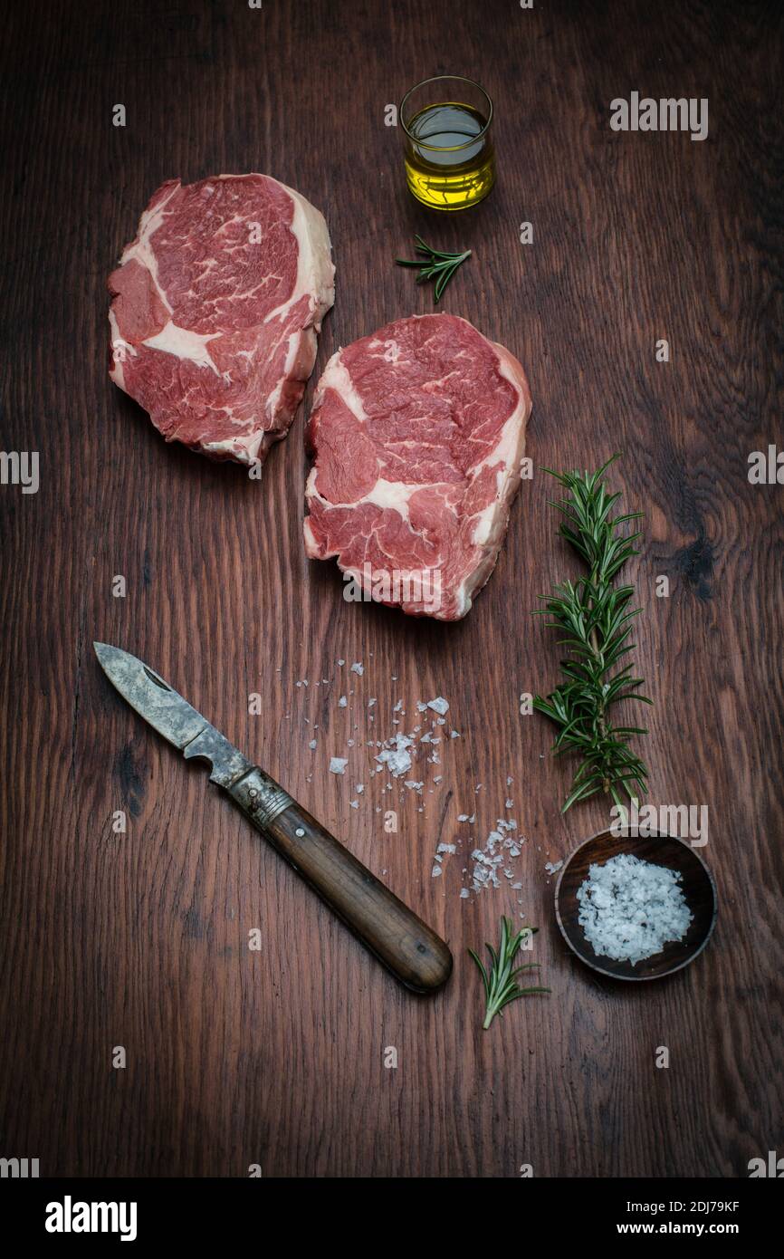 Zwei Rindersteaks garniert mit einem Rosmarinzweig, Olivenöl, Meersalzgranulat und einem Messer auf Holzgrund. Stockfoto