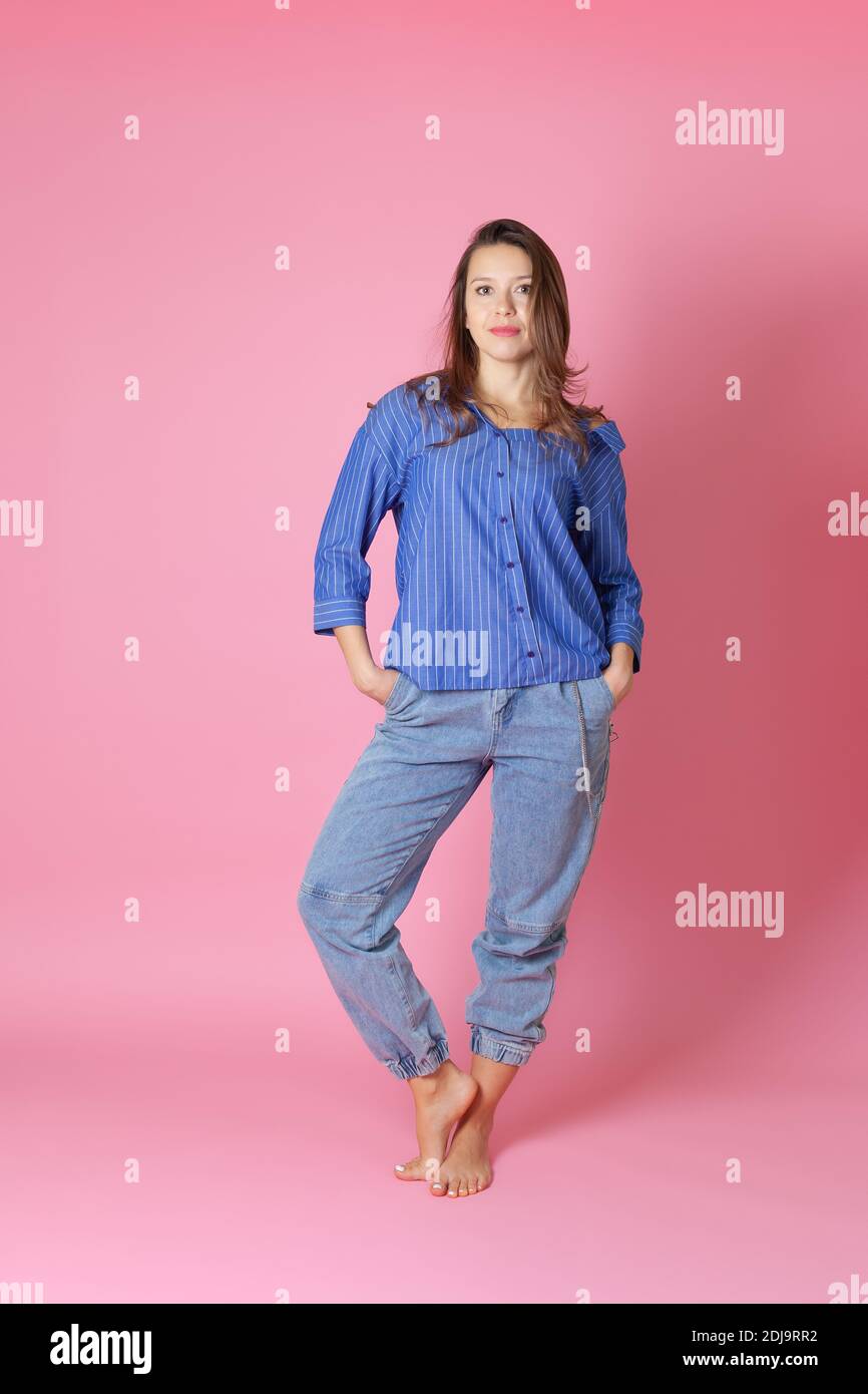 Ganzkörperportrait einer lächelnden europäischen jungen Frau in einem blauen Hemd und Jeans, die ihre Hände in den Taschen versteckt, isoliert auf einem rosa Hintergrund Stockfoto