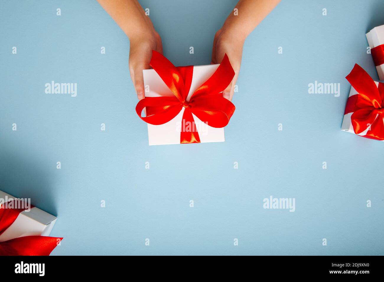 Kinderhände halten eine weiße Geschenkbox mit einer roten Schleife auf blauem Hintergrund. Urlaub Geschenk Konzept.Flat Lay Stockfoto