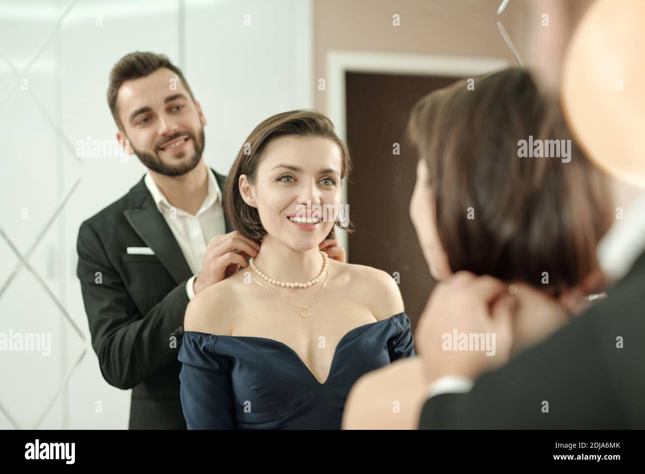 Lächelnder bärtiger Mann in Anzug trägt Halskette zu Freundin in Vor dem Spiegel, während sie sich zum Abendessen anziehen Restaurant Stockfoto