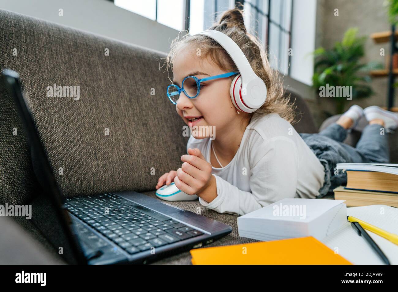 8 Jahre altes Mädchen in der Brille, das online von zu Hause aus mit einem Laptop auf dem Sofa im Zimmer liegt. Fernunterricht während der Coronavirus-Pandemie CO Stockfoto