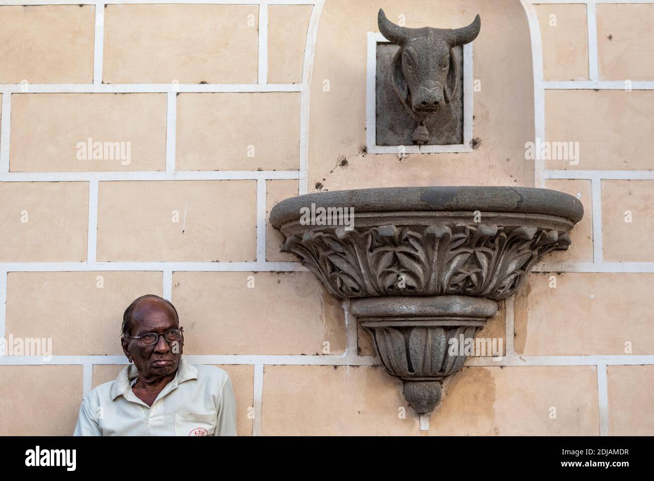 Jamnagar, Gujarat, Indien - Dezember 2018: Ehrliches Porträt eines älteren indischen Mannes, der neben einer alten Skulptur an den Wänden eines Palastes sitzt. Stockfoto