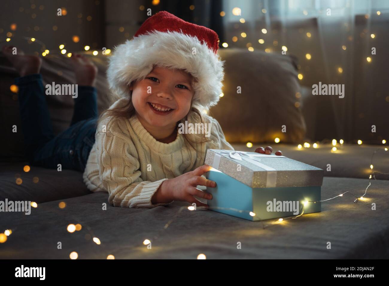 Das kleine Mädchen trägt in Santa rot Kappe öffnen magische Geschenkbox mit schönem Licht im Inneren. Magische Erfüllung von Wünschen. Weihnachtskonzept. Stockfoto