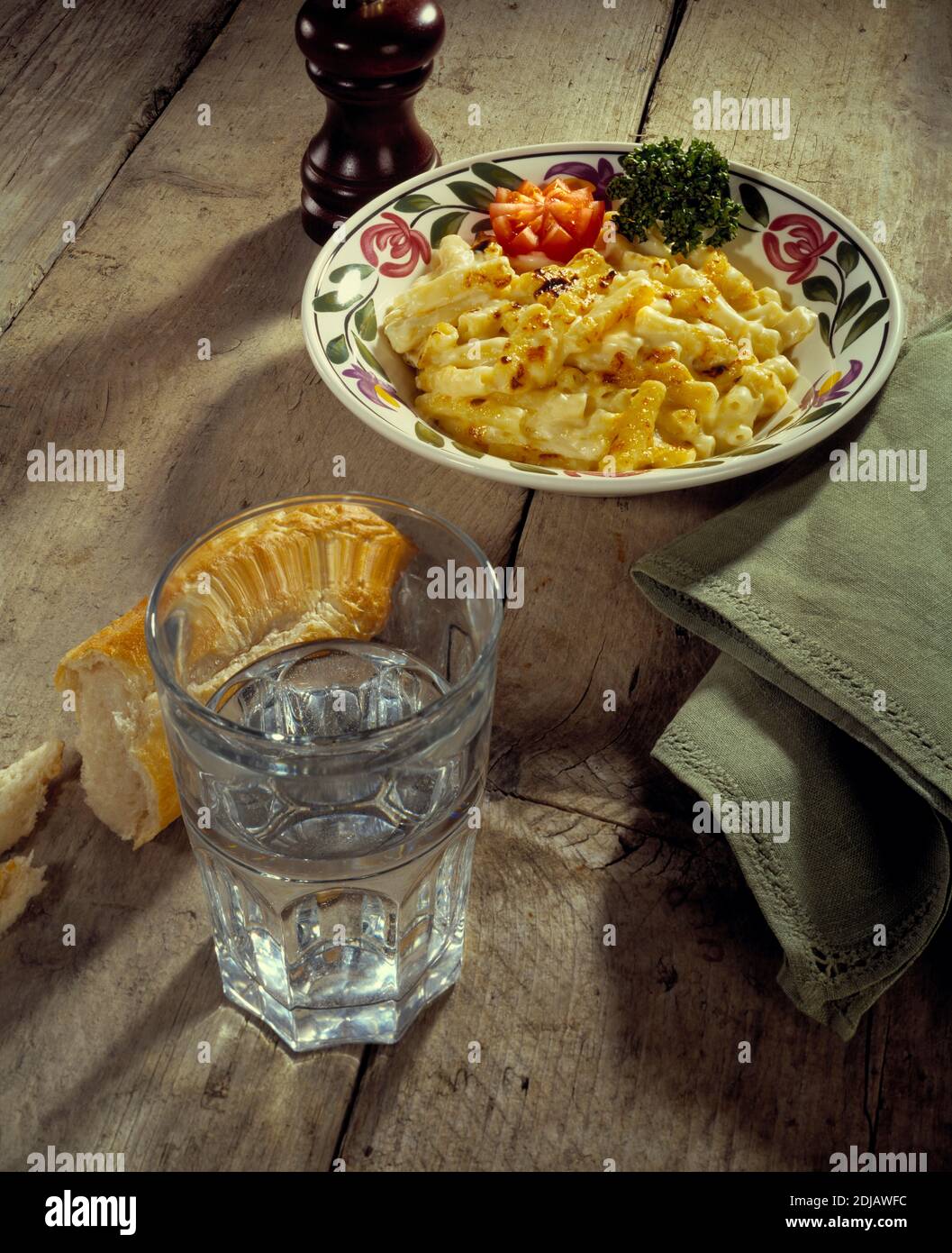 Mac n Cheese Pasta mit Wasserglas, Brot und Pfeffermühle, auf Holzhintergrund mit olivfarbener Serviette Stockfoto