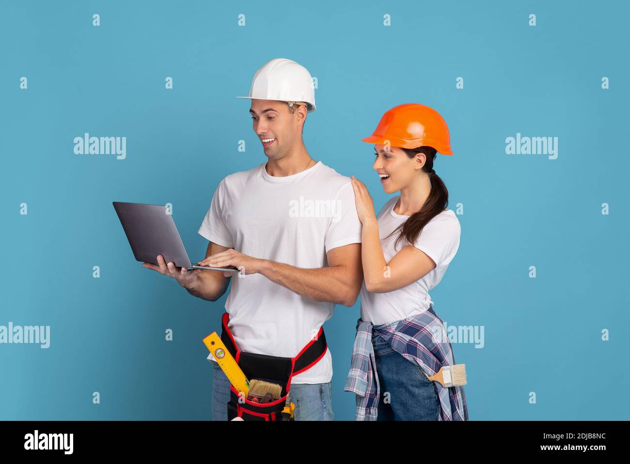 Junges Paar in Hardhats mit Laptop während Hausreparatur, blauer Hintergrund Stockfoto
