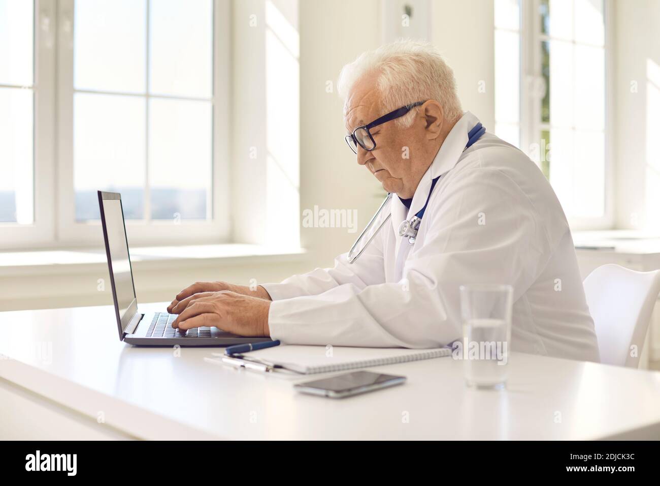 Gealterter professioneller Arzt in Uniform sitzt am Laptop und kommuniziert Mit dem Patienten online Stockfoto