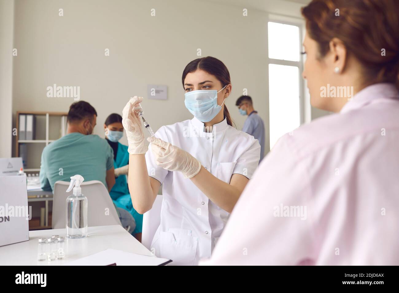 Krankenschwester in einer Maske wählt den Impfstoff in eine Spritze, während sie in einem Krankenhaus neben einem Patienten sitzt. Stockfoto