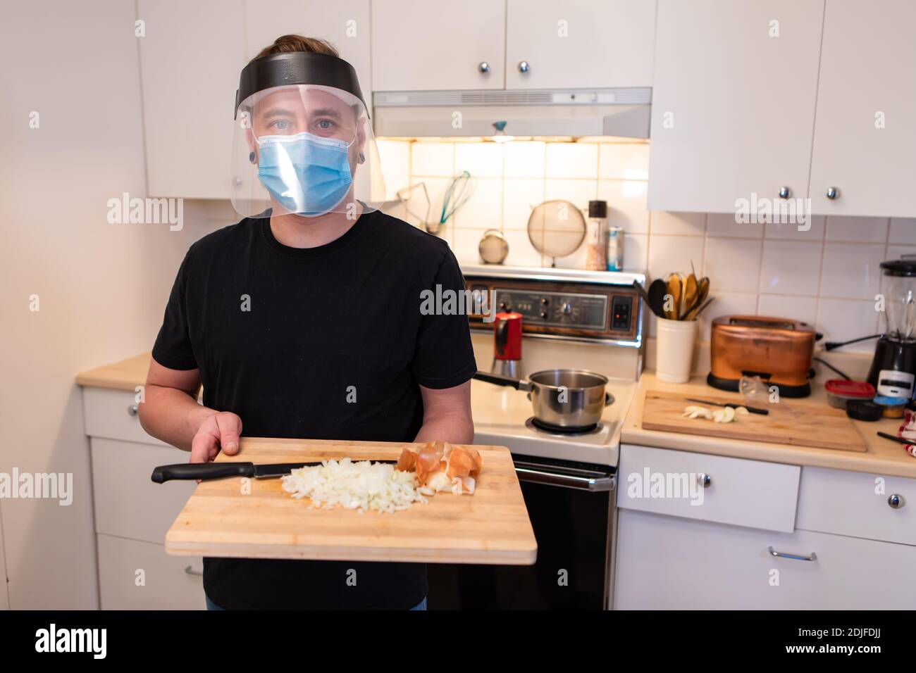 Mittlere Nahaufnahme eines jungen Mannes in einer Küche, der ein Schneidebrett mit gewürfeltem Gemüse hält, eine Covid-Schutzmaske und ein Plastikvisier trägt Stockfoto
