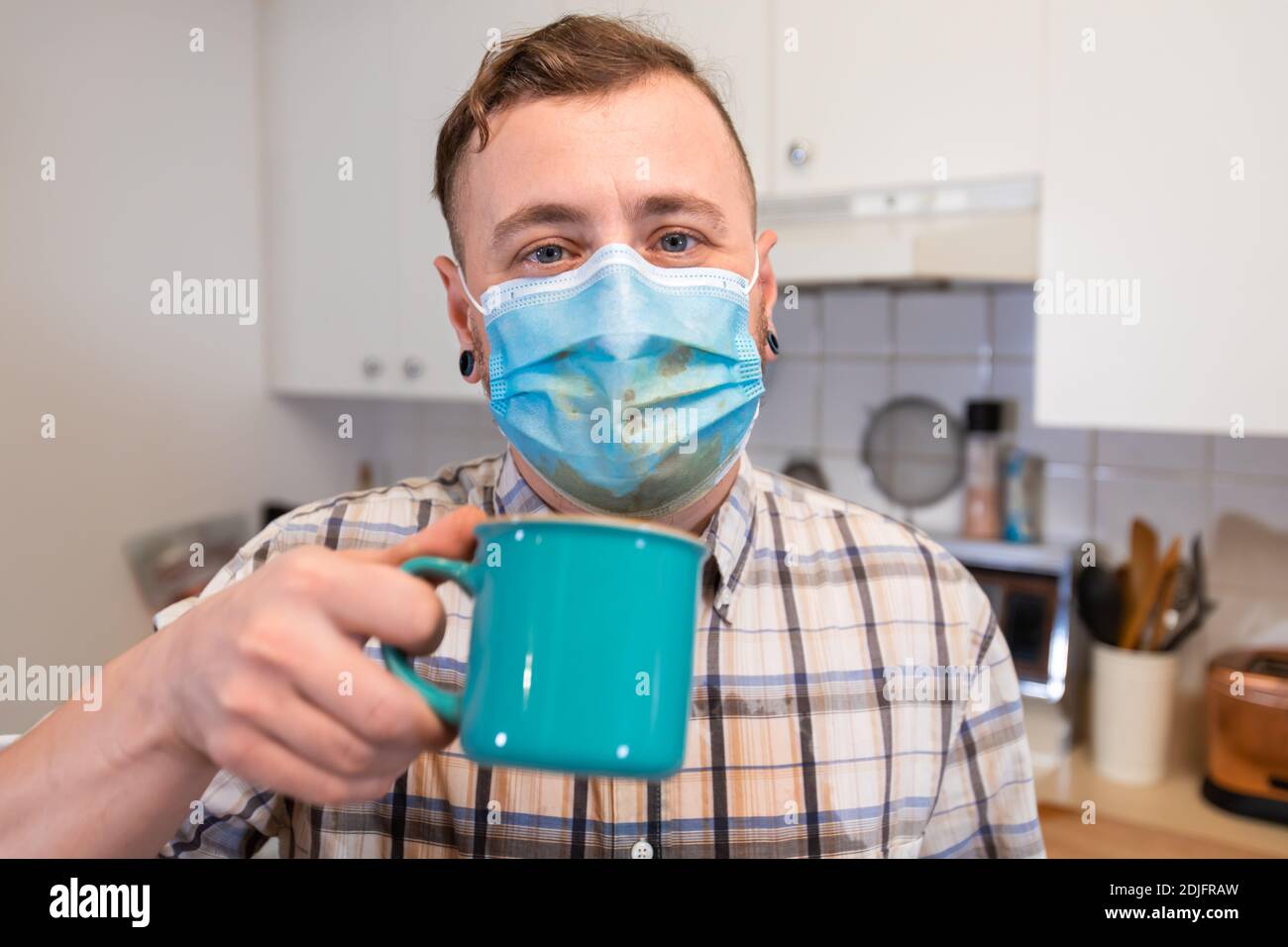 Coronavirus-Verrücktheit. Nahaufnahme eines Mannes, der offenbar versucht, seinen Kaffee mit einem covid Schutz Gesichtsmaske ein Chaos zu trinken. Ironische Stimmung. Stockfoto