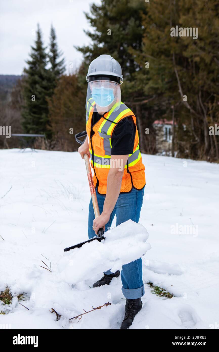 Voller Schuss eines Mannes, der eine Fluo Visibility Jacke, Helm, covid Gesichtsmaske und Kunststoff-Visier trägt, schaufelt er Schnee in einer Auffahrt und Blick auf die Kamera Stockfoto