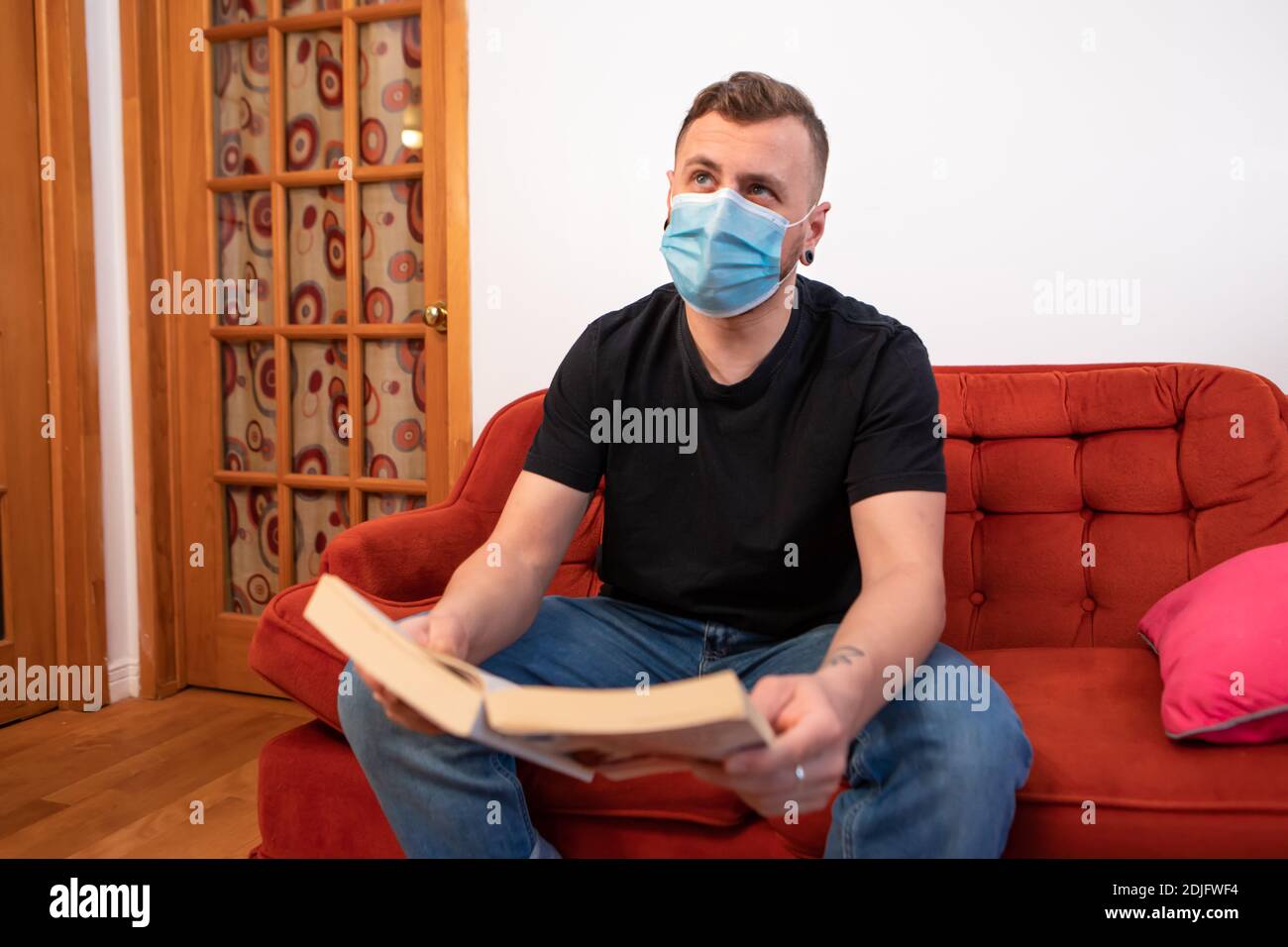 Übertriebene Covid-Prävention. Mann liest ein Buch auf einer roten Couch, trägt eine schützende Sanitär-Maske gegen covid und schaut mit lustigen Ausdruck Stockfoto