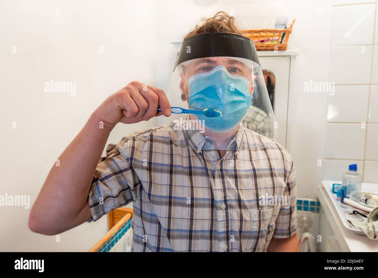 Lustige und ironische Aufnahme eines jungen Mannes in seinem Badezimmer, der versucht, seine Zähne mit und über einer covid Schutzmaske und Plastikvisier zu waschen. Stockfoto