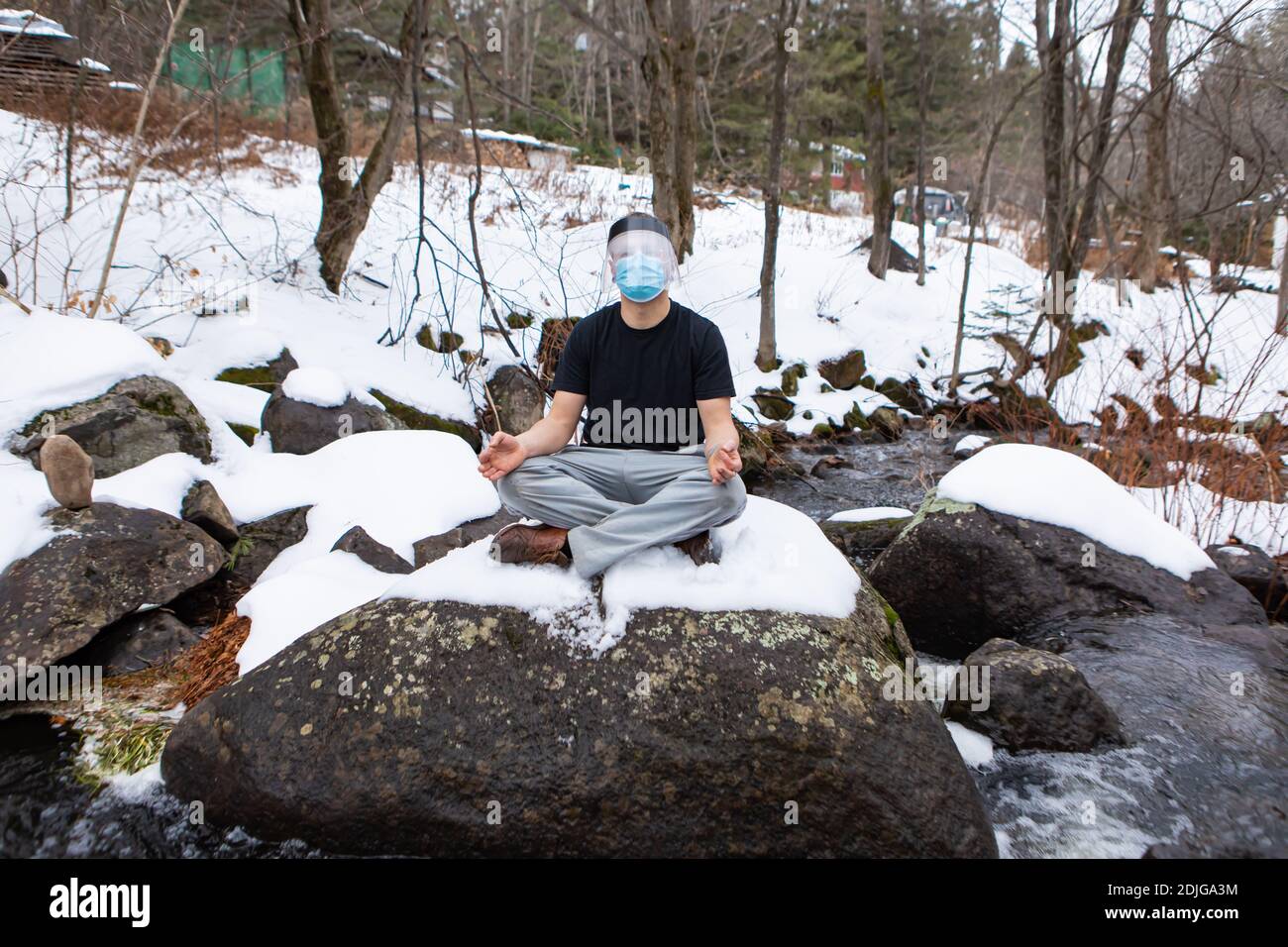 Weitwinkelaufnahme eines Mannes in einem T-Shirt, der in der Yoga-Lotusblüte auf einem großen Felsen im Schnee sitzt, eine Covid-Maske und ein Schutzvisier trägt. Stockfoto