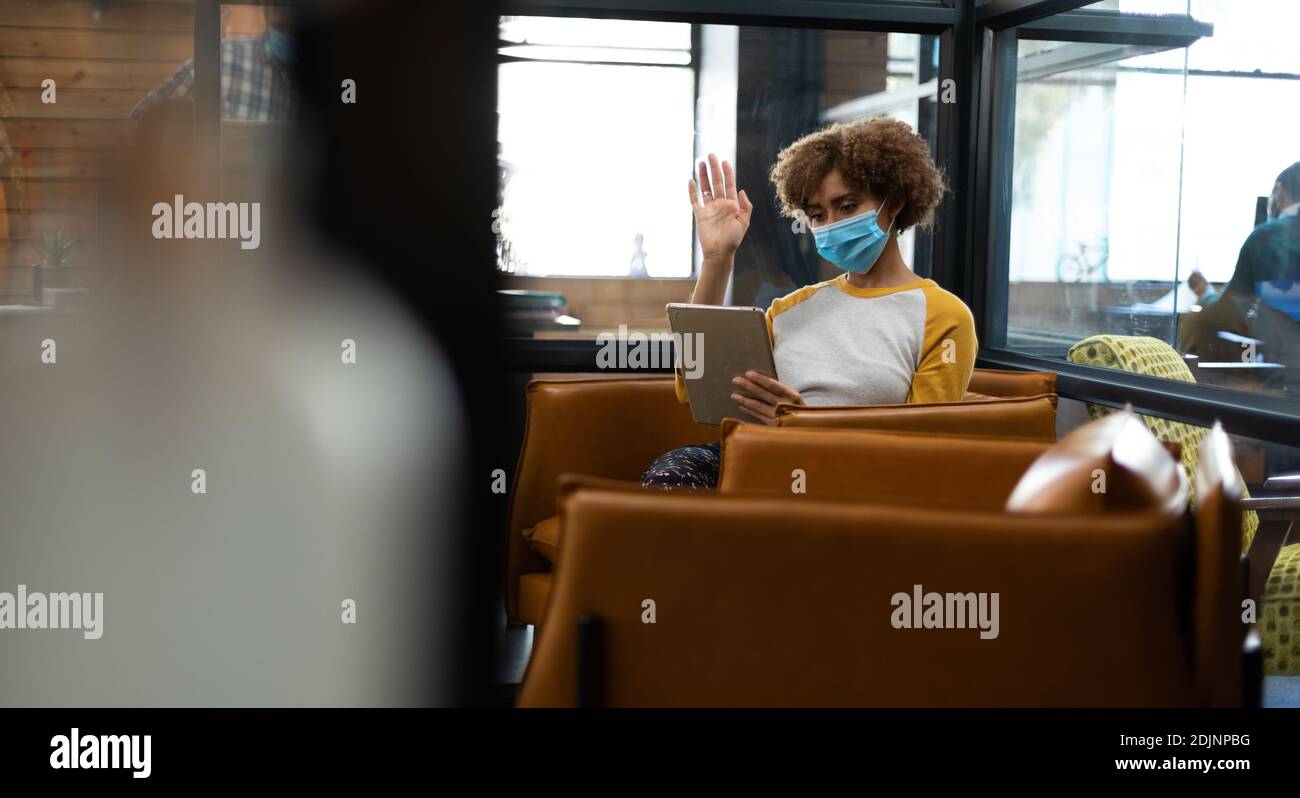 Mixed Race Frau trägt Gesichtsmaske mit Tablet und Kopfhörer Winken während eines Videotreffens Stockfoto