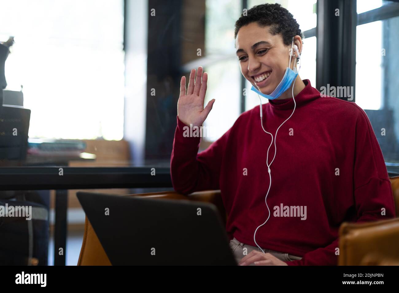 Mixed Race Frau trägt gesenkt Gesichtsmaske mit Laptop und Ohrhörer, die während einer Videokonferenz winken Stockfoto