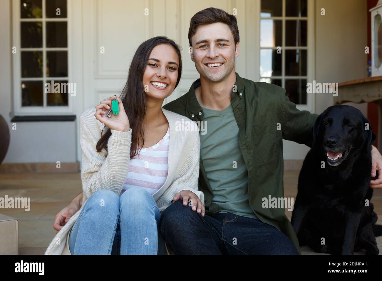 Lächelndes kaukasisches Paar, das Schlüssel vor dem Haus mit Hund zeigt Stockfoto