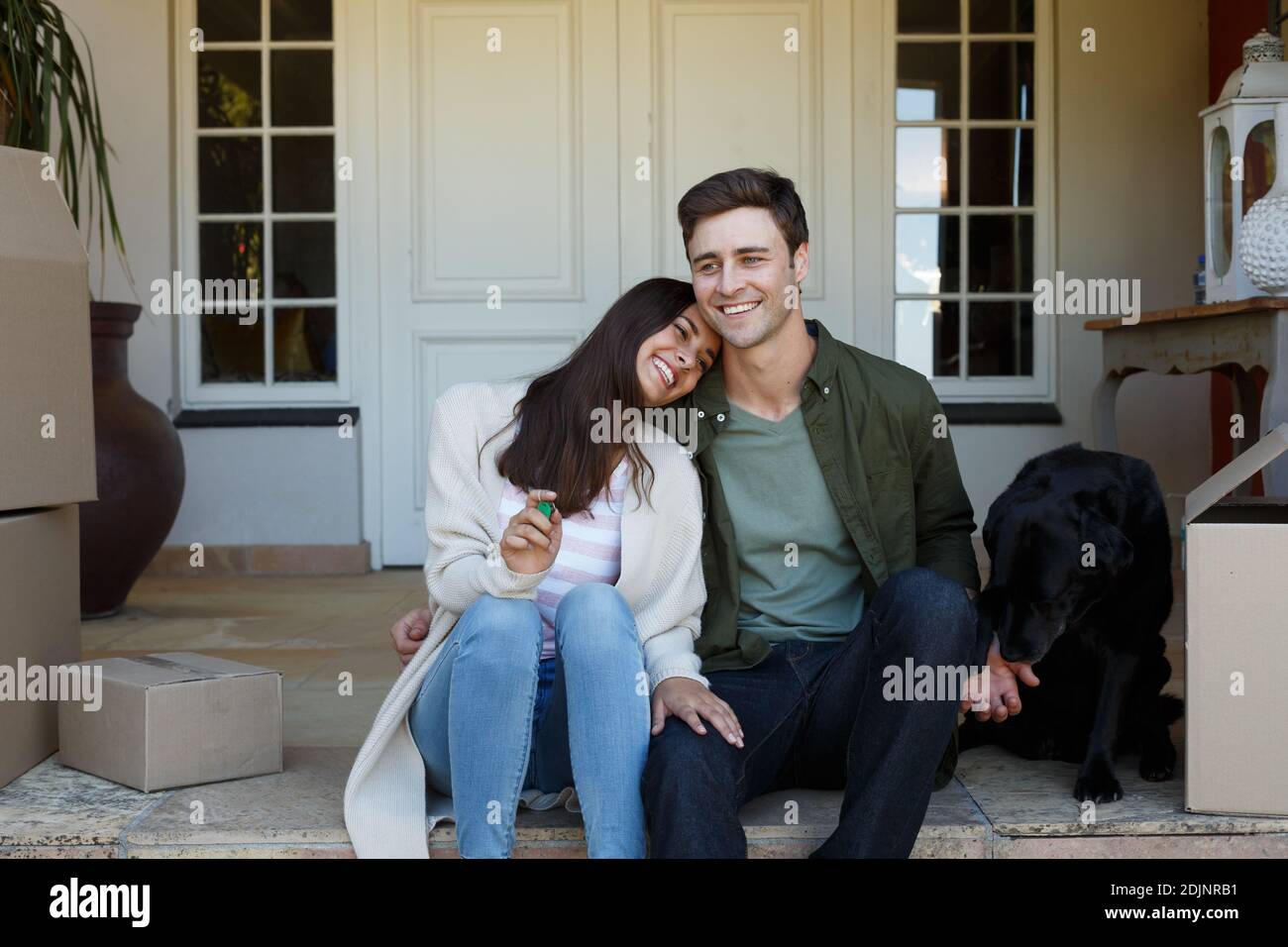 Lächelndes kaukasisches Paar mit Schlüsseln, das vor dem Haus mit Hund sitzt Stockfoto