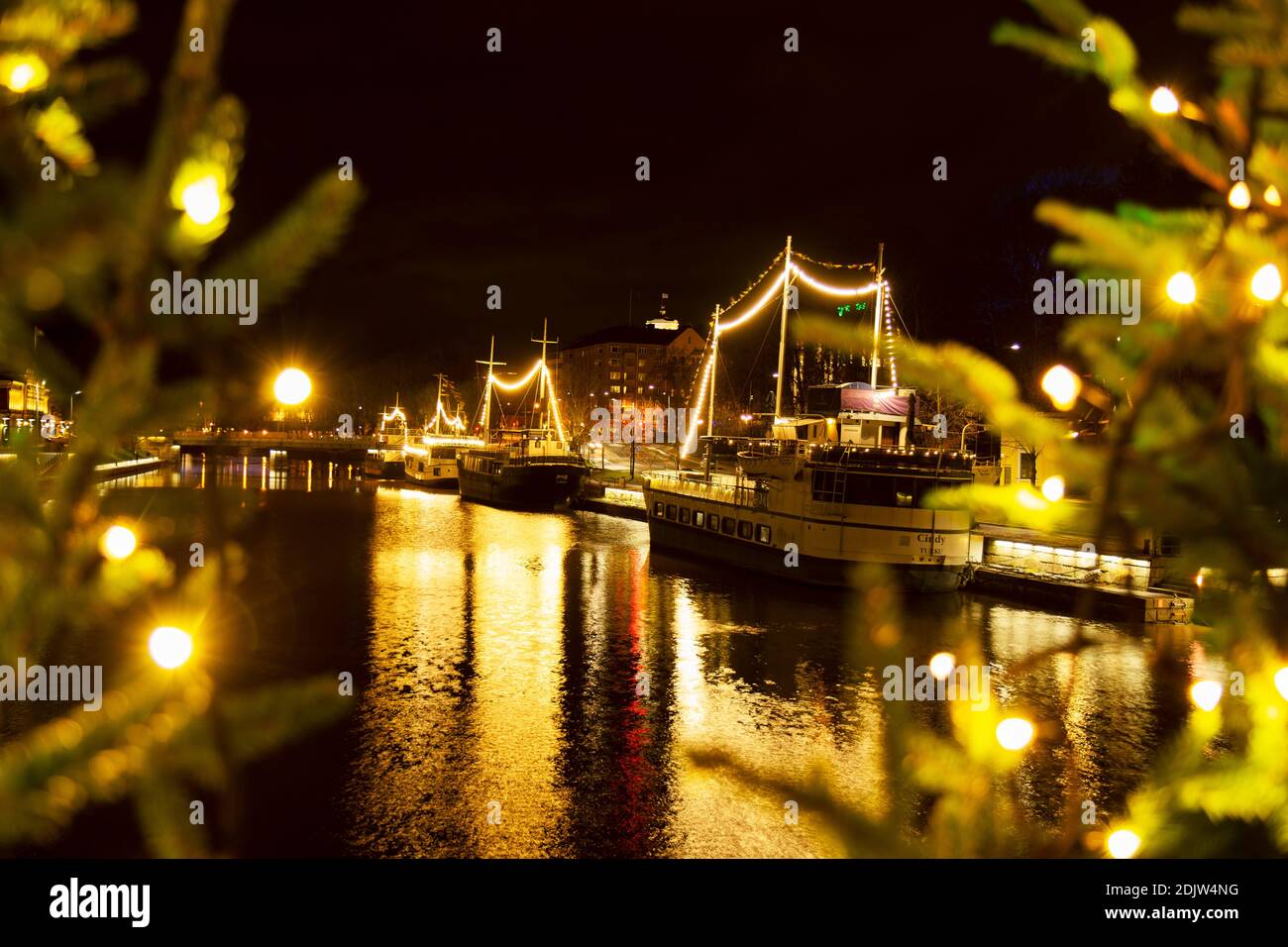 Die Restaurantboote liegen am Aura-Fluss mit wunderschöner nächtlicher Beleuchtung. Verschwommener Vordergrund mit beleuchteten Christbaumzweigen gefüllt. Stockfoto