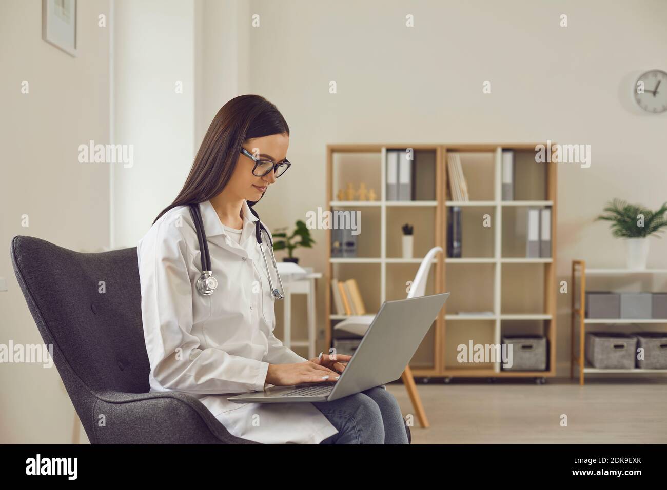 Ärztin, die mit Laptop arbeitet, während sie im Stuhl im Hintergrund des Büros sitzt. Stockfoto