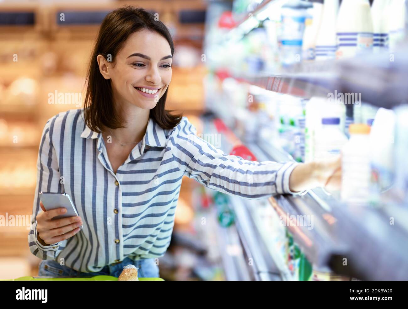 Frau hält Smartphone im Laden stehend und nimmt Milch Stockfoto