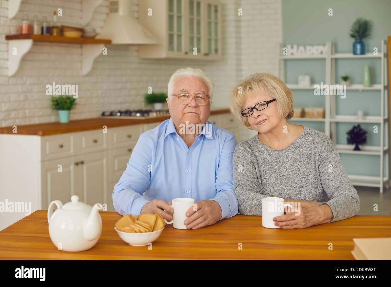 Seniorenpaar sitzt in der Küche und schaut auf die Kamera und trinkt Tee oder Kaffee mit Keksen. Stockfoto