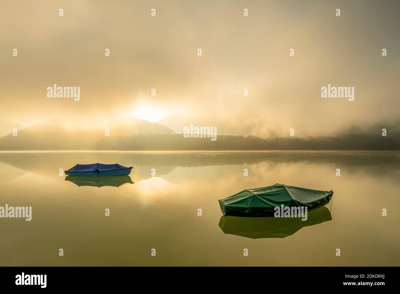 Ruderboot mit Plane auf dem Sylvensteinspeichersee im Karwendelgebirge. Im Hintergrund Nebel bei Sonnenaufgang mit einer einzigartigen Lichtatmosphäre und ruhiges Wasser. Stockfoto