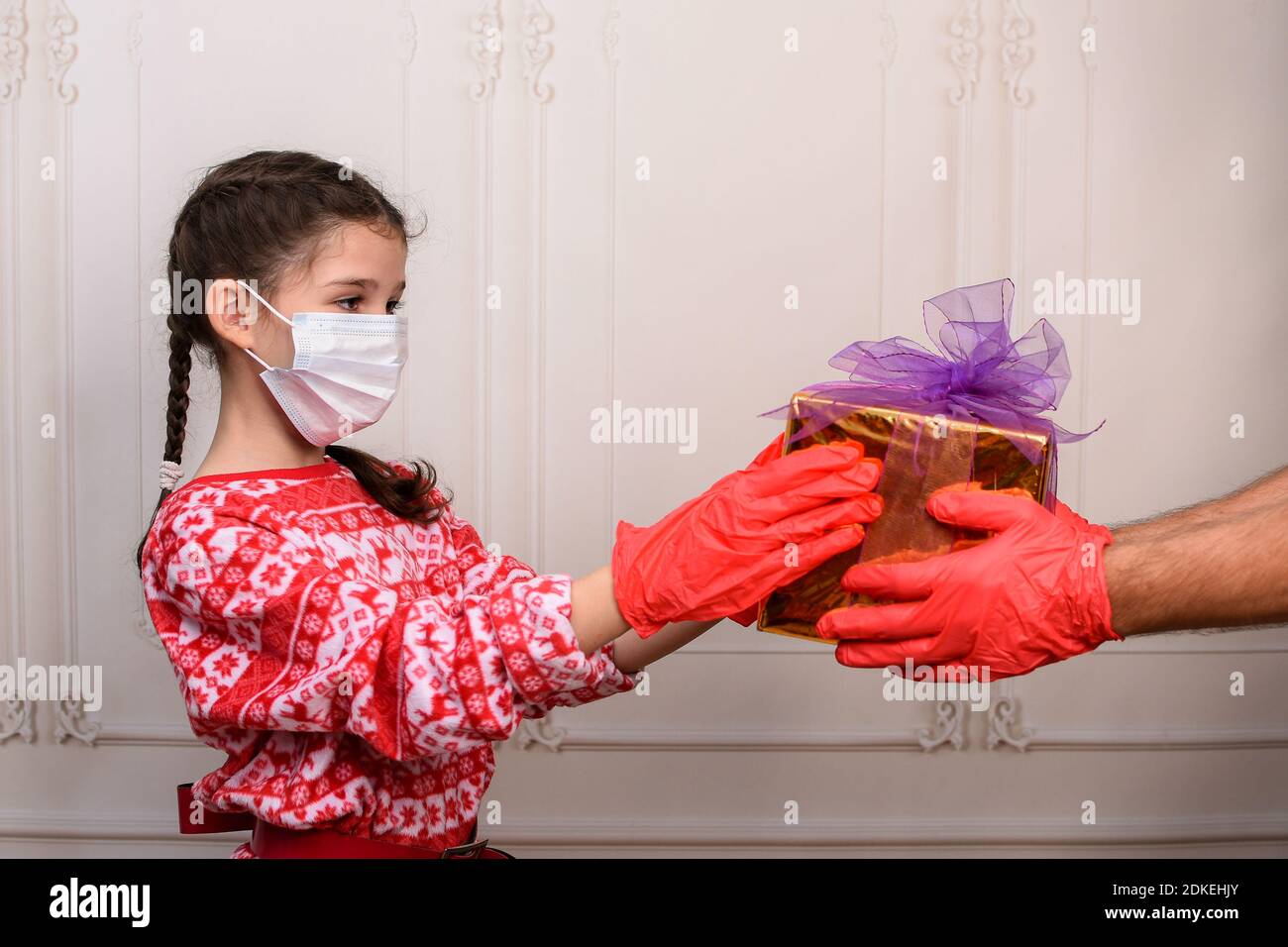 Kindheit und Menschen, das Konzept von Weihnachten während der Epidemie. Mädchen in einer Maske und Handschuhe erhält ein Geschenk. Stockfoto