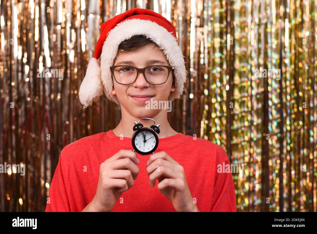 Porträt eines Jungen mit Brille und in einem Weihnachtsmann Hut mit einer Uhr, die die Zeit auf einem goldenen Hintergrund anzeigt. Weihnachtsdekoration. Zwölf Uhr Stockfoto