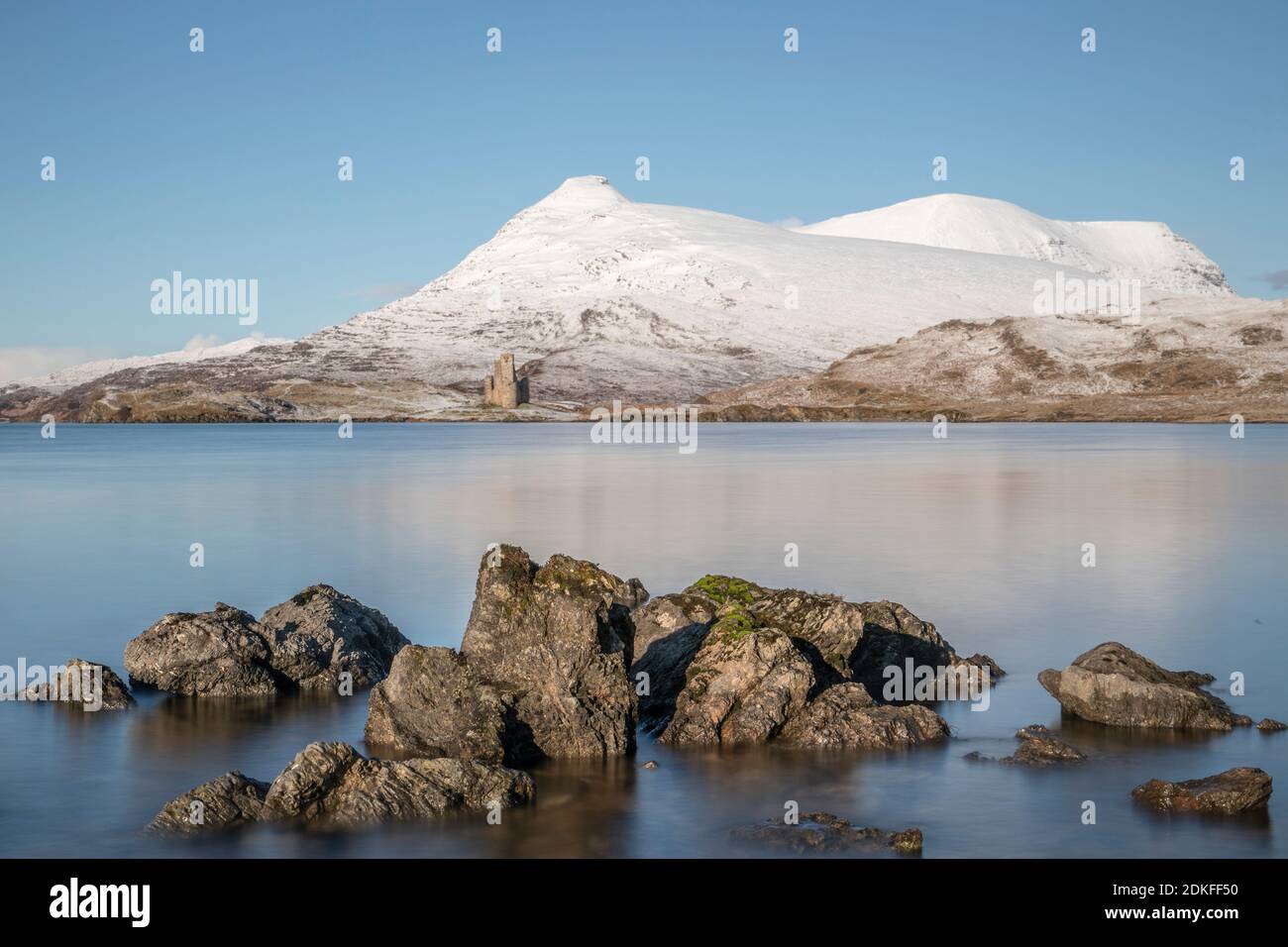 Blick über Steine am Ufer des winterlichen Loch Assynt, Schottland, Ruine von Ardvreck Castle mit schneebedeckten Bergen im Hintergrund Stockfoto