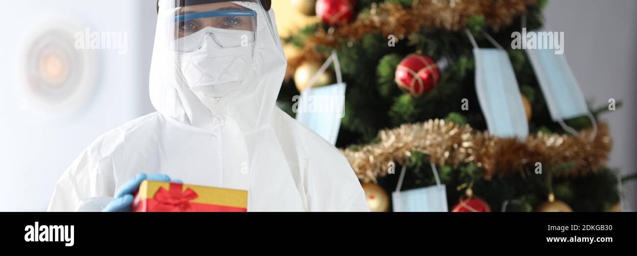 Arzt im Schutzanzug steht neben Weihnachtsbaum und Hält Geschenk Stockfoto