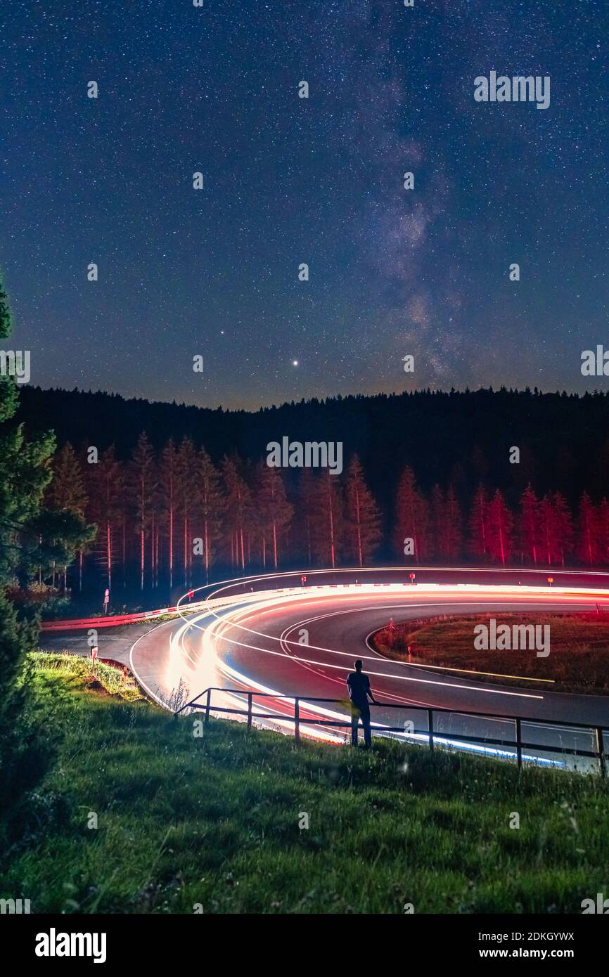 Milkyway, Langzeitbelichtung eines Mannes, der an einer Geländer vor einer Straße steht, während Autos vorbeifahren und Cartrail-Licht verlassen. Stockfoto