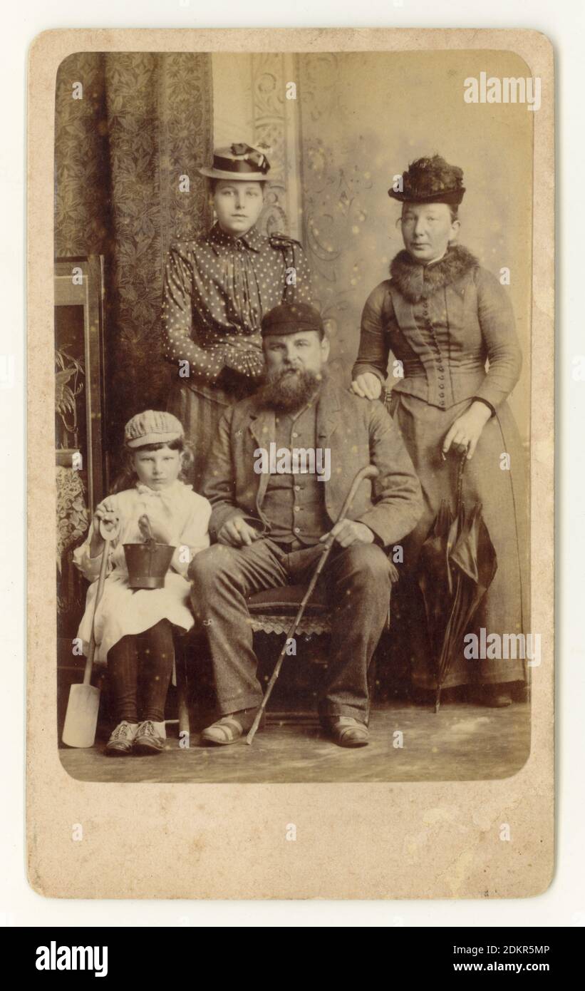 Original charaktervolles viktorianisches CDV (Carte de Visite) Erinnerungsfoto einer Arbeiterfamilie mit Schwiegermutter im Urlaub im Badeort Skegness, Lincolnshire, England, U.K. um 1888. Das Kind, das einen Eimer und Spaten hält, trägt eine merkwürdige Mischung aus Mädchenkleid und Knabenmütze und ist angesichts der viktorianischen Gewohnheit, Jungen in Mädchenkleidung zu kleiden, wahrscheinlich ein Junge. Er sieht nicht sehr glücklich aus und lässt sein Foto offenbar unter Zwang machen. Stockfoto