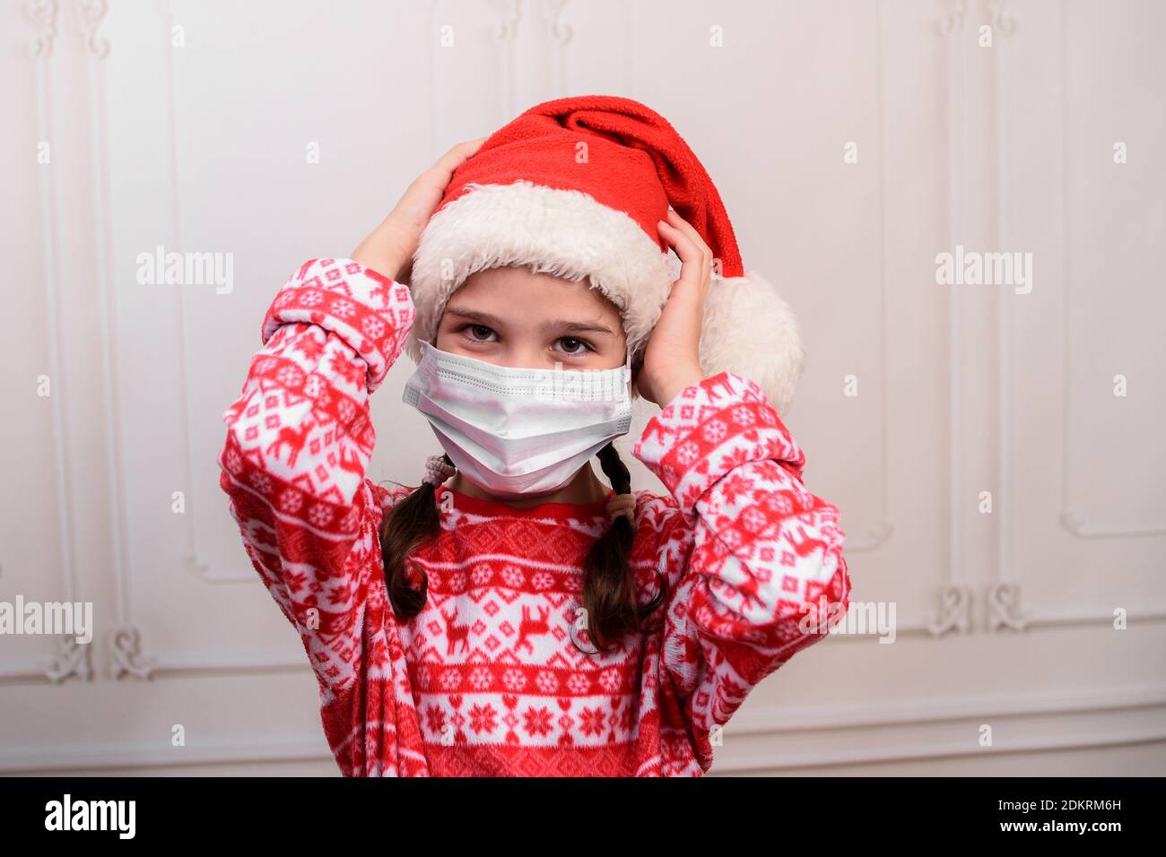 Lustige kleine Mädchen trägt einen Weihnachtsmann Hut und eine medizinische Maske. Kind Mädchen mit glücklichem Ausdruck am weihnachtstag. Stockfoto