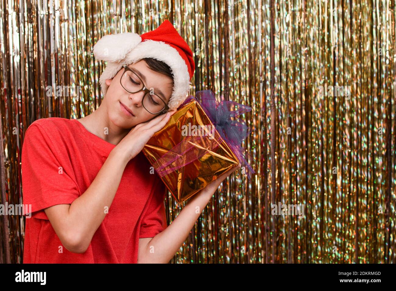 Junge mit einem Geschenk für das neue Jahr auf einem goldenen Hintergrund. Junge Teenager mit Brille und in einem Weihnachtsmann Hut mit . Weihnachtsdekoration. Stockfoto