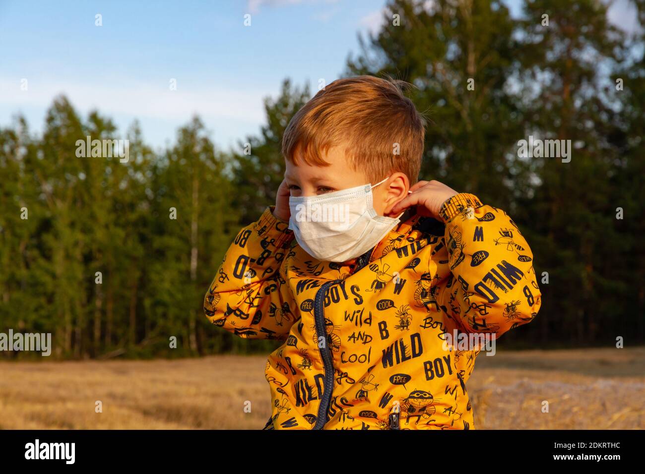 Ein Junge in einer gelben Jacke zieht einen Arzt an Maske an einem hellen sonnigen Tag Stockfoto