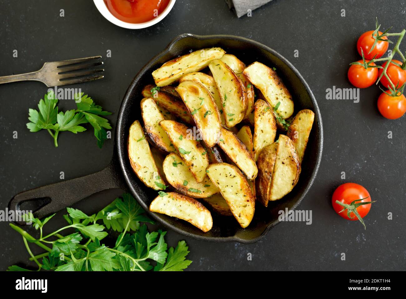 Hausgemachte Bratkartoffelkeile mit Gewürzen in Bratpfanne auf schwarzem Stein Hintergrund. Draufsicht, flach liegend Stockfoto