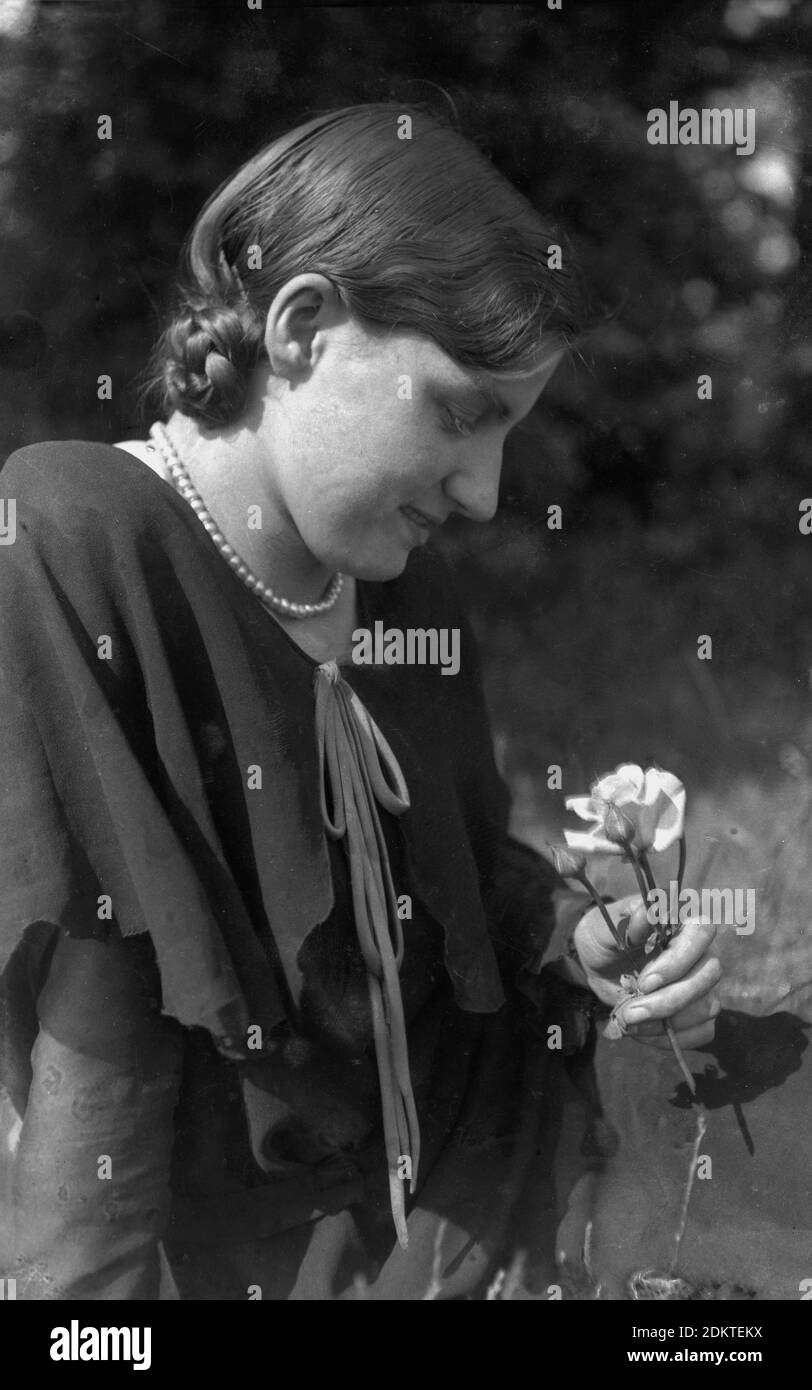 1930er Jahre, historische, junge Dame trägt ein Seidentop und Perlenkette sitzen draußen Blick auf eine Blume. Stockfoto