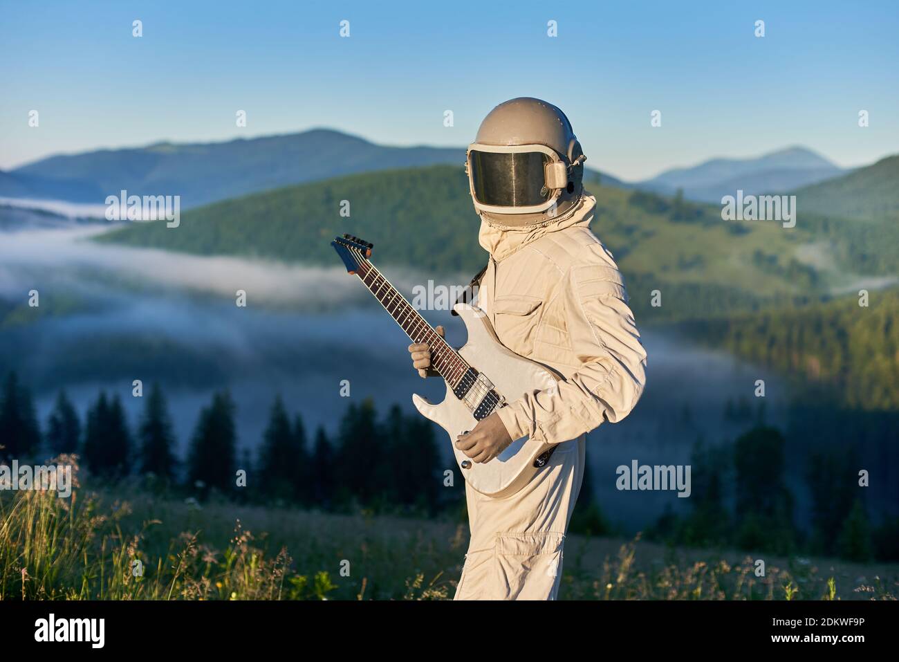 Portrait des Raumfahrers trägt weißen Raum Anzug und Helm mit weißen Gitarre, auf einer grünen sonnigen Lichtung, fantastische Landschaft des Morgens nebligen Hügeln auf dem Hintergrund Stockfoto