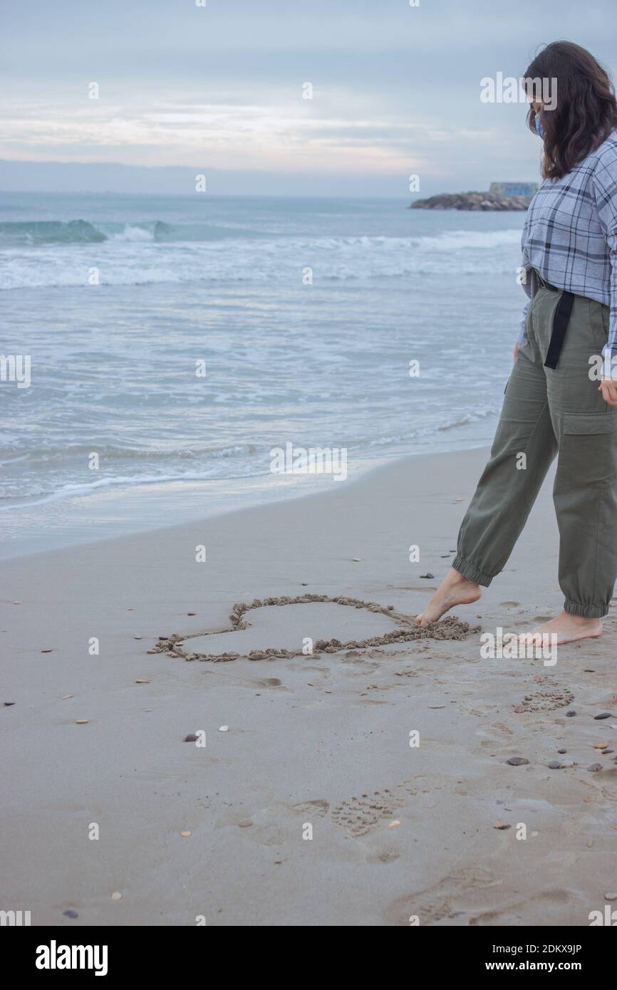 Vertikales konzeptionelles Bild. Erkennbare Frau zieht am 14. Februar mit dem Fuß ein Herz auf den Sand. Valentinstag 2021. Stockfoto