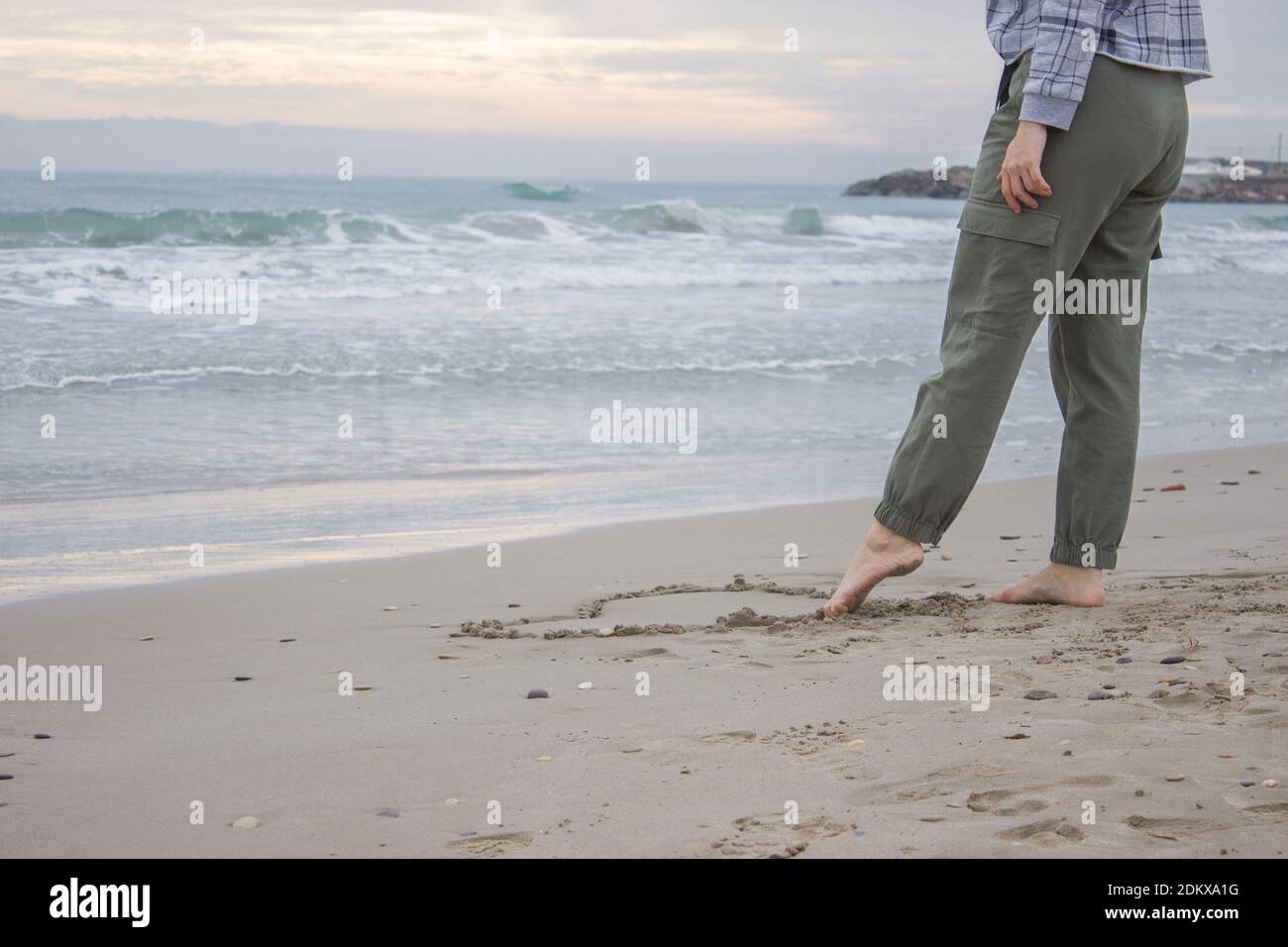 Horizontales konzeptionelles Bild mit Ozeanhintergrund. Eine unkenntliche Frau zieht am 14. Februar mit dem Fuß ein Herz auf den Sand. Valentinstag Stockfoto