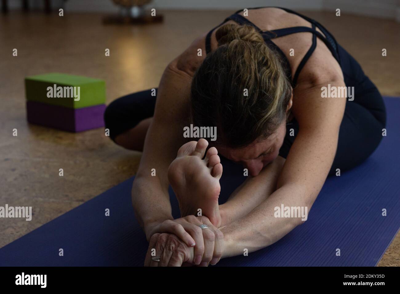 Flexible Frau auf Kopf bis Knie Yoga Pose mit Requisiten auf der Seite im Studio. Weibliche Yogi praktiziert janu sirsasana mit Händen zusammen wickeln Fuß Stockfoto