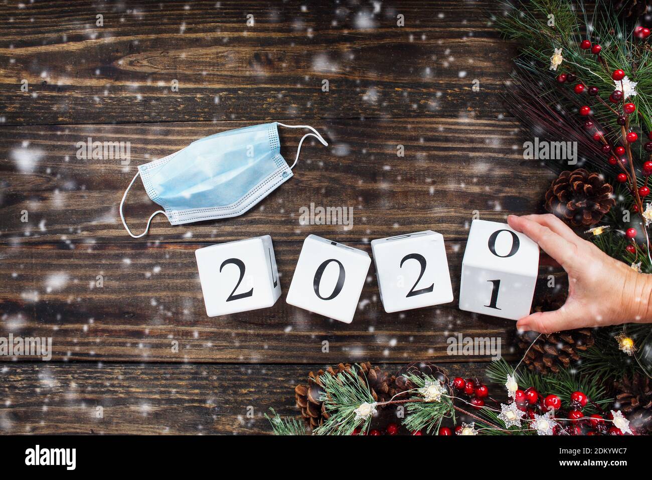 Frau Hand kippt Neujahr 2020 Holz Kalender Blöcke zu 2021. Medizinische Maske, Weihnachtsbaumlichter, Dekorationen, Kiefernzweige, rote Winterberrie Stockfoto