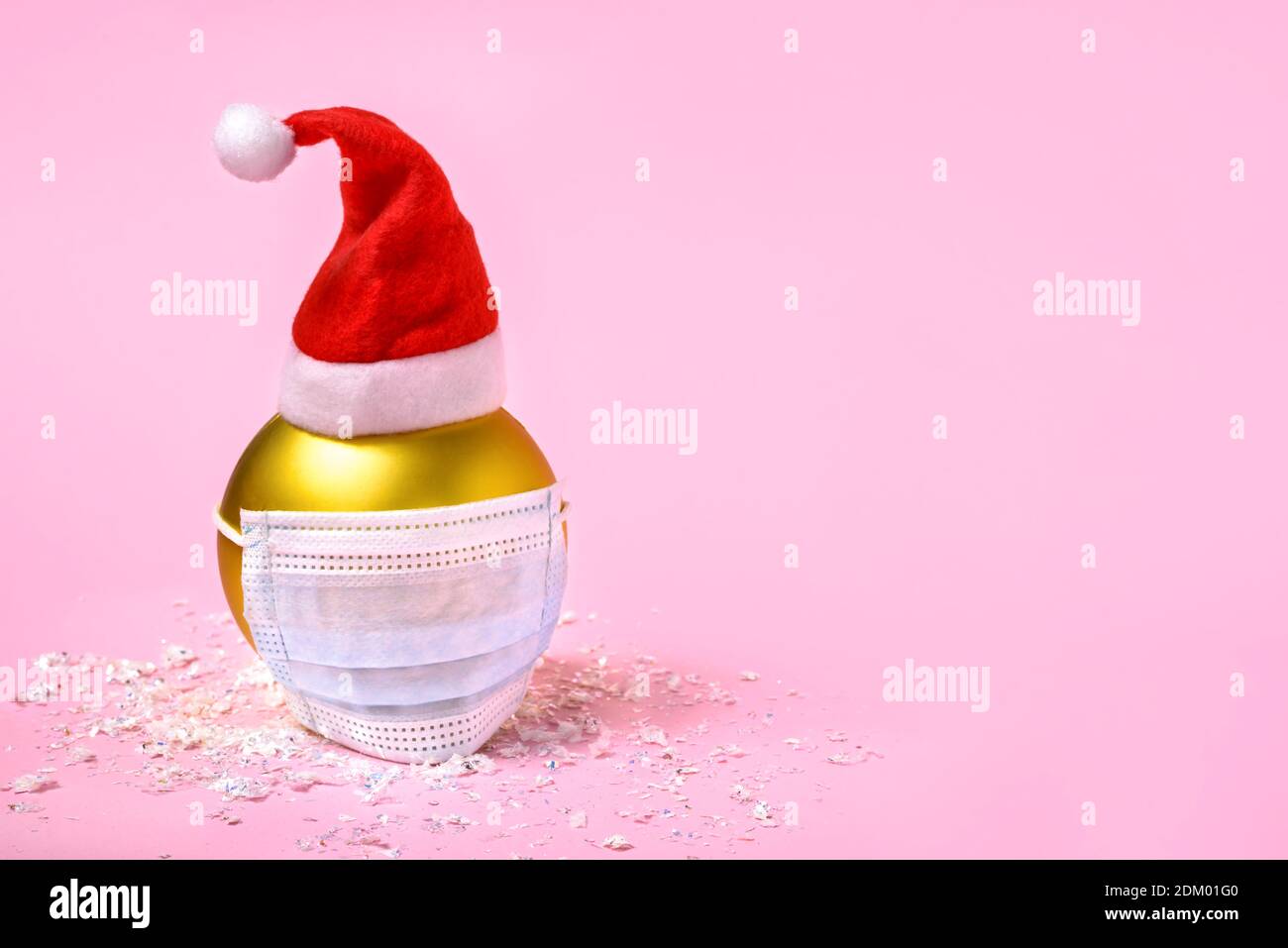 Goldene Weihnachtskugel mit blauer chirurgischer Gesichtsmaske und Weihnachtsmann-Hut auf rosa Hintergrund mit Platz für Text. Stockfoto