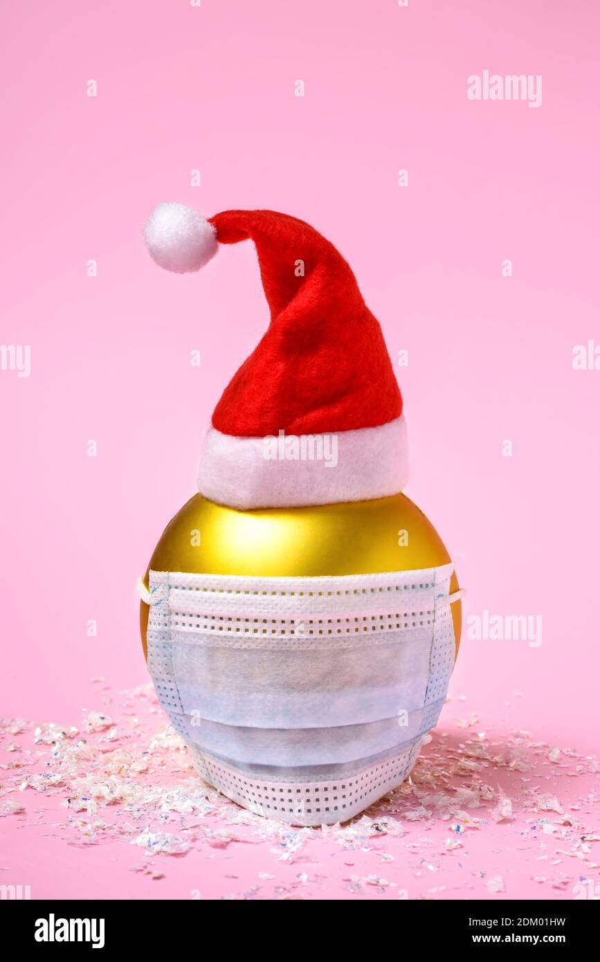 Goldene Weihnachtskugel mit blauer chirurgischer Gesichtsmaske und Weihnachtsmann Hut auf rosa Hintergrund. Stockfoto