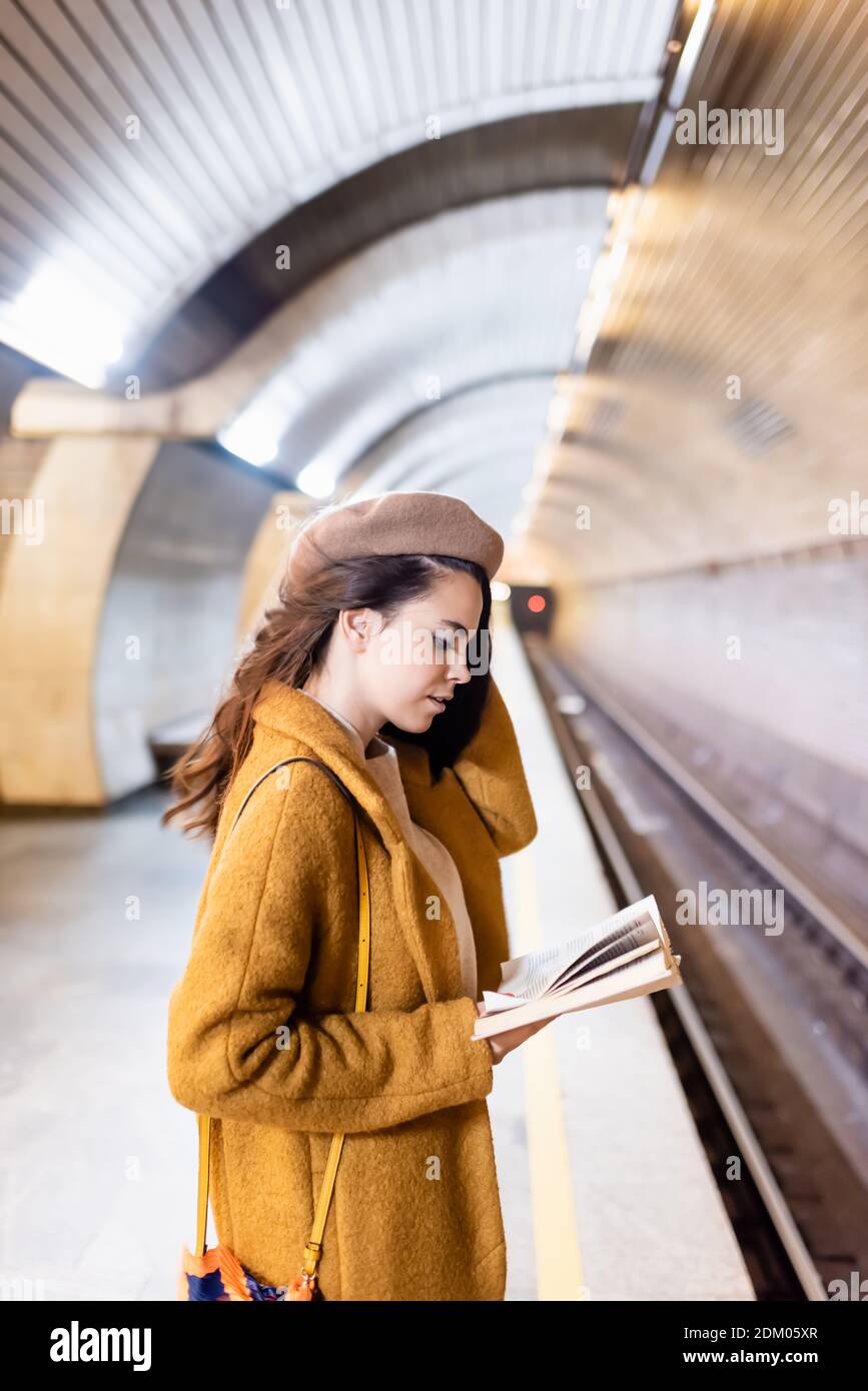 Junge Frau im Mantel und Baskenmütze liest Buch über U-Bahn Plattform Stockfoto