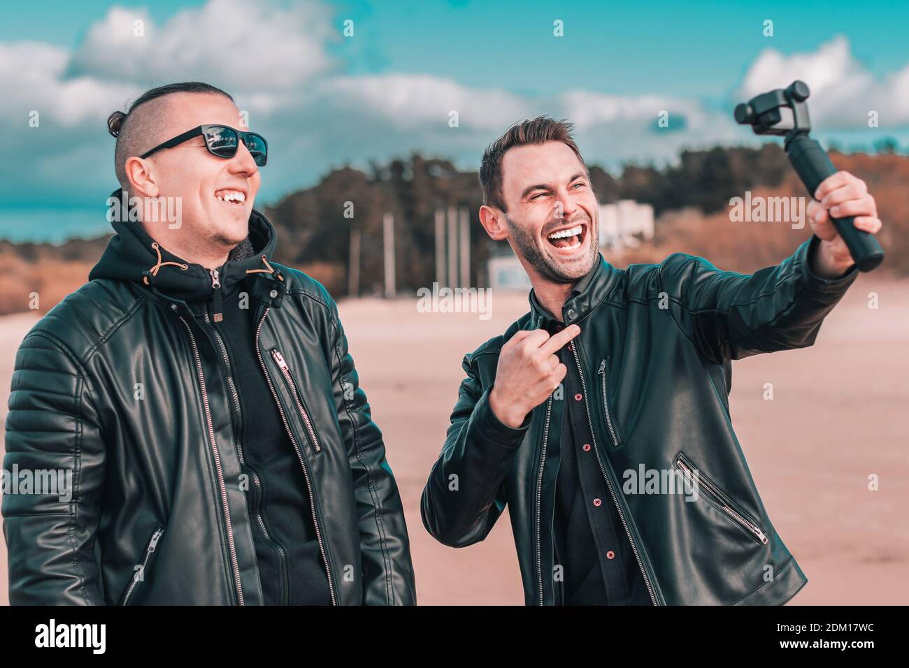 Zwei gutaussehende lächelnde Freunde machen Selfie mit Action-Kamera mit Gimbal Stabilisator am Strand. Jugendliche Männer in schwarzen Kleidern Spaß haben, indem Sie Fotos Stockfoto