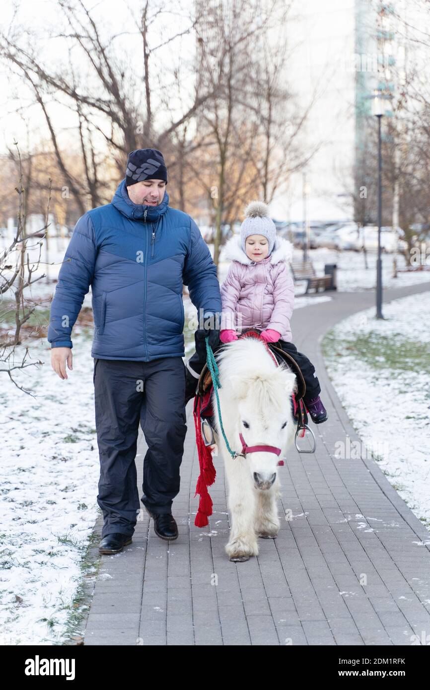 Papa und kleines Mädchen in einem gestrickten Hut und warm Jacke Reiten Sie ein Pony in einem Stadtpark auf einem Sonniger Wintertag Stockfoto